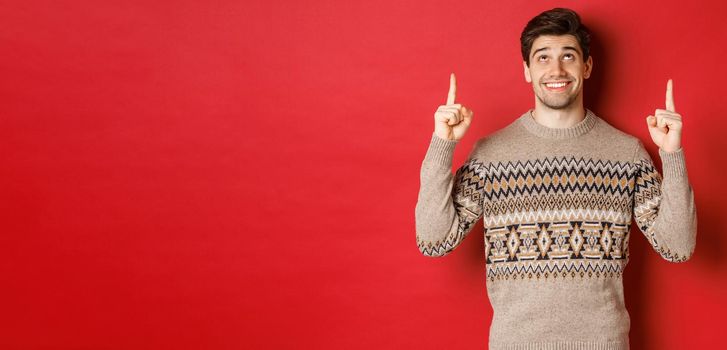 Portrait of dreamy handsome guy thinking about christmas holidays, wearing winter sweater, pointing and looking up at new year promo offer, standing over red background.