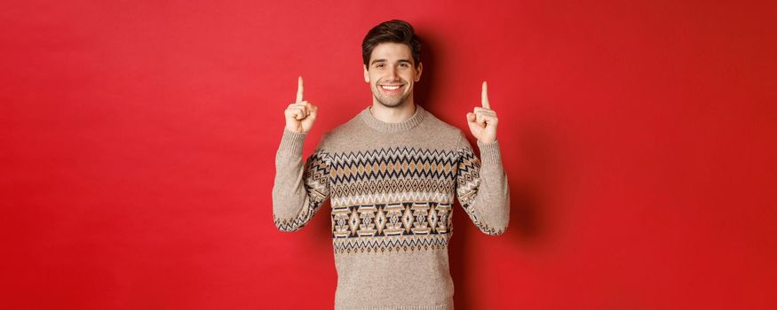 Portrait of attractive guy in christmas sweater pointing fingers up, smiling and showing winter holidays advertisement, celebrating new year over red background.