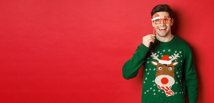 Portrait of handsome smiling man in christmas sweater and party glasses, looking surprised and happy, celebrating new year and having fun, standing against red background.