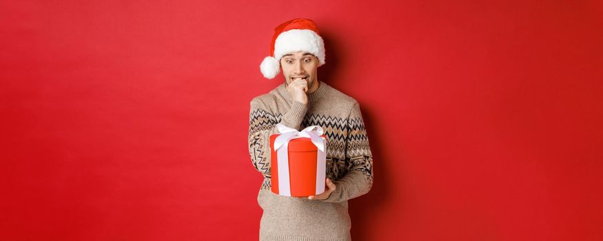 Image of worried guy in santa hat and sweater, looking indecisive at christmas gift, standing against red background.