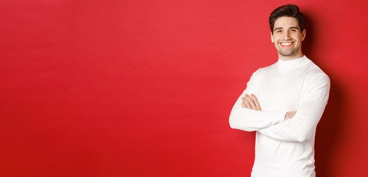 Concept of winter holidays, christmas and lifestyle. Portrait of confident attractive guy in white sweater, smiling with hands crossed on chest, standing over red background.