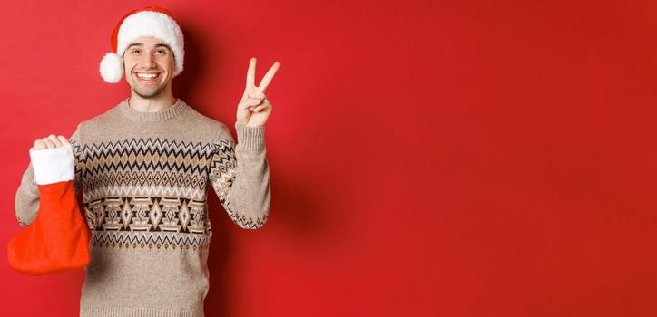 Concept of winter holidays, new year and celebration. Image of happy smiling man in santa hat and sweater, showing peace sign and a christmas stocking bag with gifts, red background.