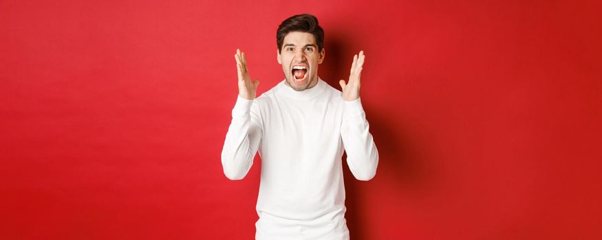 Image of frustrated and angry man in white sweater, shouting in rage, being mad at someone, standing over red background.