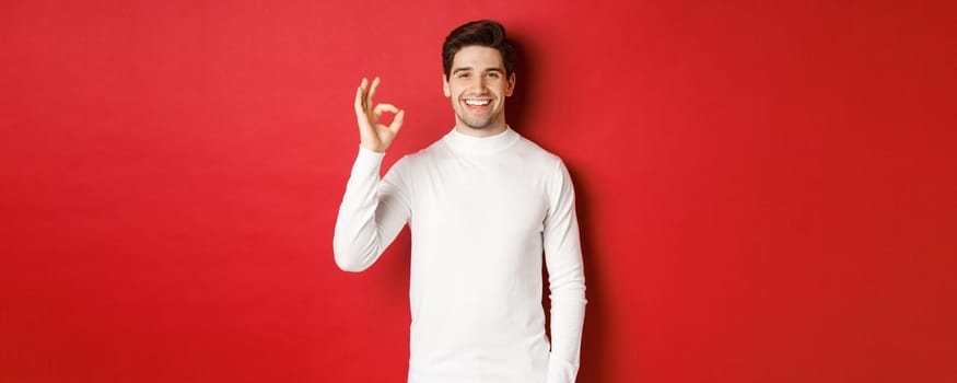 Concept of winter holidays, christmas and lifestyle. Happy handsome man in white sweater showing okay sign, smiling pleased, recommending shop, standing over red background.