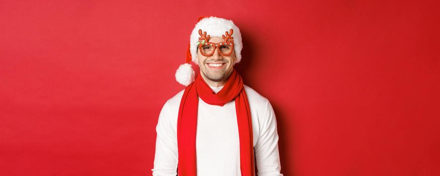 Concept of christmas, winter holidays and celebration. Cheerful handsome guy in new year party glasses and smiling, standing over red background.