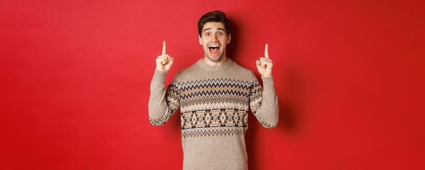 Image of handsome excited man, feeling happy about christmas holidays, pointing fingers up and looking cheerful, showing advertisement, standing in sweater over red background.