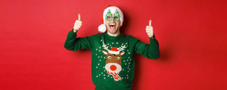 Portrait of super happy young man in party glasses, santa hat and sweater, showing thumbs-up in approval, recommending new year promo offer, standing over red background.