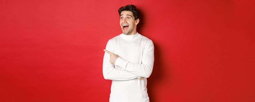Concept of winter holidays. Image of amazed handsome man reacting to big christmas announcement, looking and pointing left excited, standing over red background.