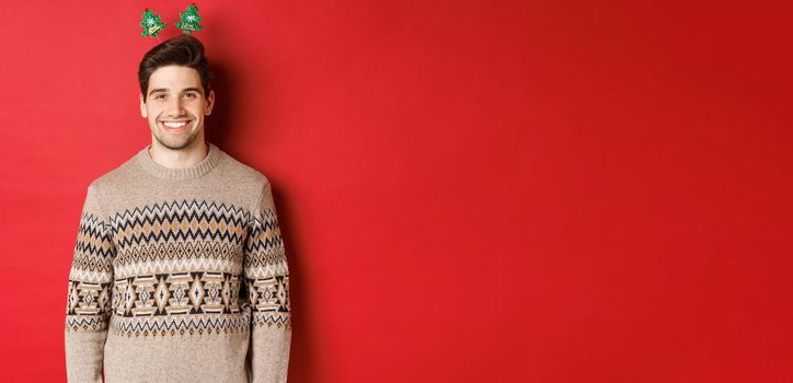 Concept of winter holidays, christmas and celebration. Handsome bearded guy in sweater wishing happy new year, smiling at camera, standing over red background.