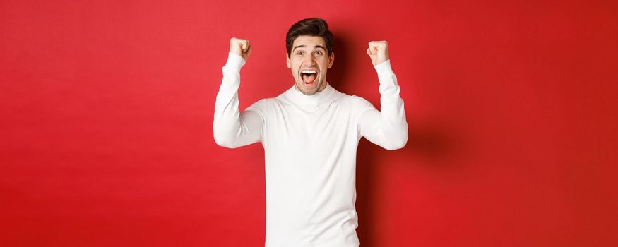 Portrait of excited lucky man in white sweater, raising hands up and triumphing, celebrating new year, standing happy against red background.
