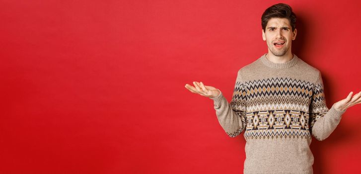 Portrait of confused and disappointed handsome guy, complaining about christmas, spread hands sideways and frowning displeased, standing in xmas sweater over red background.