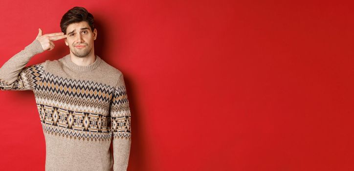 Portrait of distressed and annoyed handsome man, wearing christmas sweater, making finger gun sign near head and shooting himself, standing over red background.