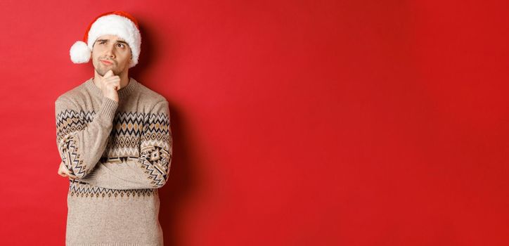 Image of attractive man in christmas sweater and santa hat, thinking about new year gifts, looking at upper left corner thoughtful, standing over red background.