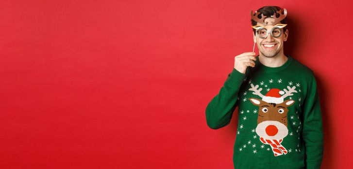 Handsome happy man in christmas sweater, holding party mask and smiling, enjoying new year celebration, standing over red background.