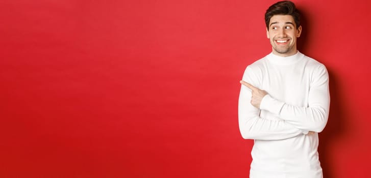 Concept of winter holidays, christmas and lifestyle. Excited handsome man in white sweater, smiling, looking and pointing finger left at promo offer, standing over red background.