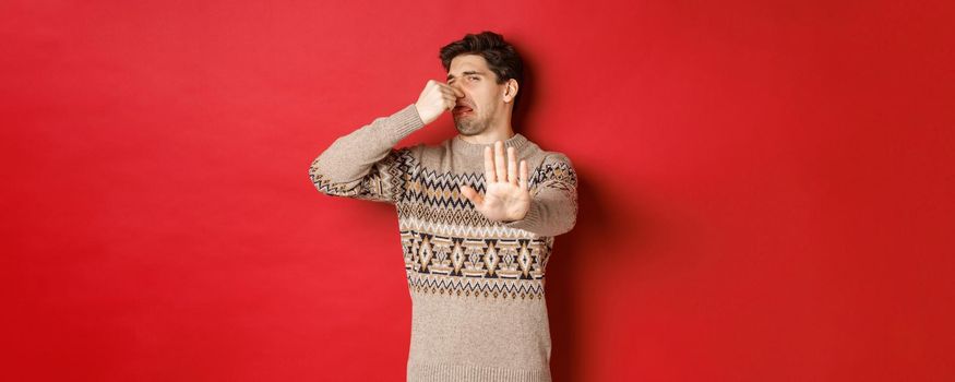 Image of disgusted handsome man in winter sweater, rejecting something with bad smell, shut nose and raising hand to decline, standing over red background.