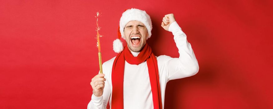 Concept of christmas, winter holidays and celebration. Handsome man celebrating new year and having fun, holding sparkler and smiling, wearing santa hat, standing over red background.