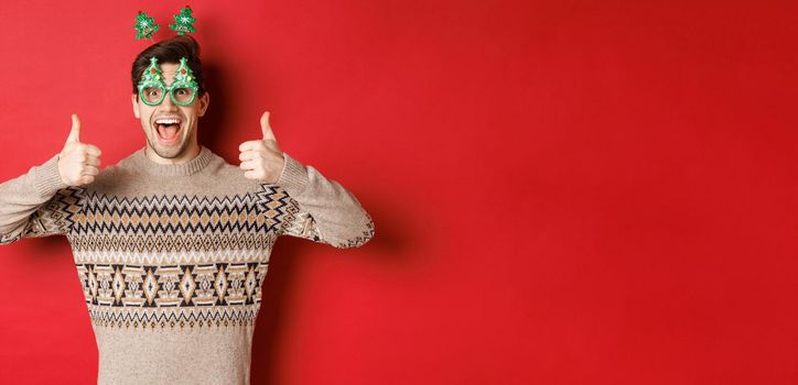 Image of excited handsome guy in party glasses and christmas sweater, showing thumbs-up, praising great new year celebration, standing over red background.