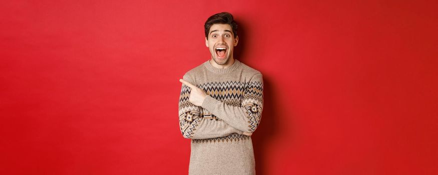 Portrait of amazed handsome guy, wearing christmas sweater, pointing finger at upper left corner logo and looking excited, showing awesome new year promo offer, standing over red background.