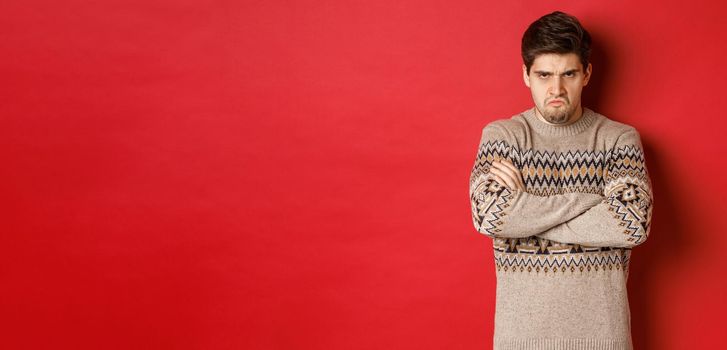 Image of angry and offedned guy in christmas sweater, feeling mad, cross arms on chest and sulking, standing over red background.