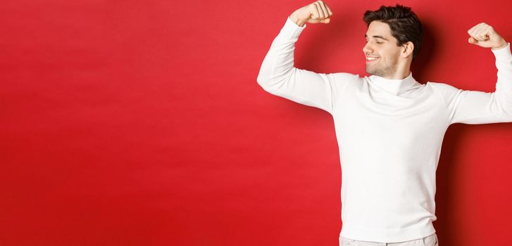 Portrait of smiling handsome man in white sweater, flexing biceps and bragging with strength, show-off strong muscles after workout, standing over red background.