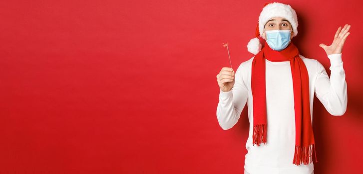 Concept of covid-19, christmas and holidays during pandemic. Cheerful handsome man in medical mask and santa hat, celebrating new year with sparkler, looking excited, red background.