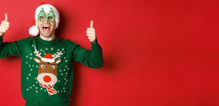 Portrait of super happy young man in party glasses, santa hat and sweater, showing thumbs-up in approval, recommending new year promo offer, standing over red background.