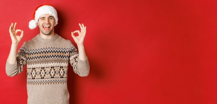 Image of joyful attractive guy in sweater and santa hat, wishing merry christmas, showing okay signs and winking at camera, celebrating new year, standing over red background.