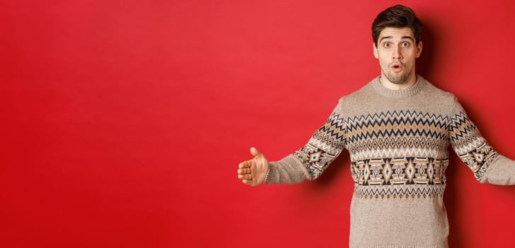 Image of amazed handsome man showing size of christmas present, spread hands sideways and shaping something large, standing over red background.