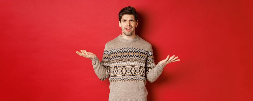Portrait of confused and disappointed handsome guy, complaining about christmas, spread hands sideways and frowning displeased, standing in xmas sweater over red background.