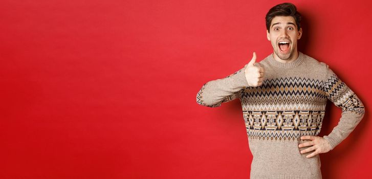 Portrait of excited, attractive man in christmas sweater, showing thumbs-up and looking amazed, recommending good thing, advertising christmas related product.