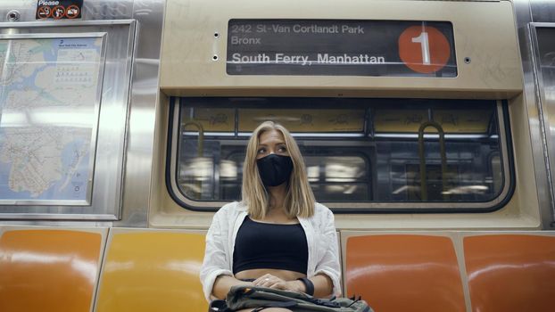 Woman in a black medical face mask to avoid the spread of coronavirus who is sitting alone in a modern subway car. Girl with cellphone in a surgical mask is keeping social distance on a metro train.