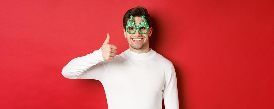 Concept of christmas, winter holidays and celebration. Close-up of handsome smiling man, wearing party glasses and white sweater, showing thumbs-up, recommending new year promo.