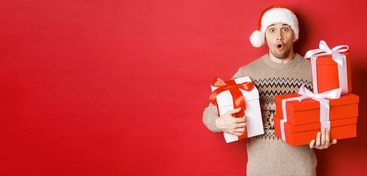 Concept of winter holidays, new year and celebration. Image of surprised handsome guy in sweater and santa hat, receiving gifts, holding boxes with presents and looking amazed.