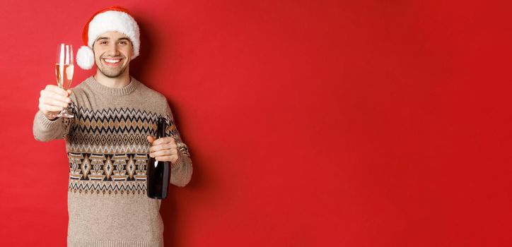 Concept of winter holidays, new year and celebration. Portrait of handsome man in santa hat and sweater, holding champagne, raising glass and saying cheers on christmas party.