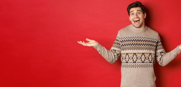 Portrait of happy and surprised, handsome caucasian guy, wearing christmas sweater, spread hands sideways and looking clueless, standing over red background.