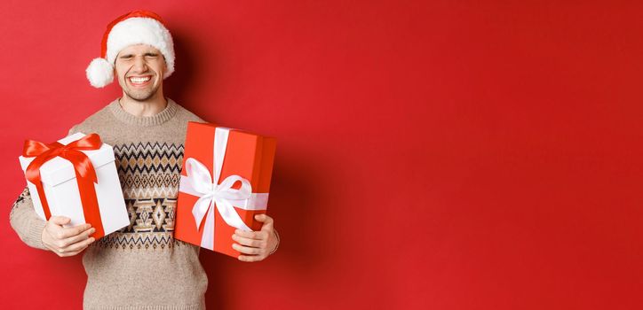 Concept of winter holidays, new year and celebration. Image of happy and excited young man likes presents, holding gifts and smiling, wearing santa hat and christmas sweater.