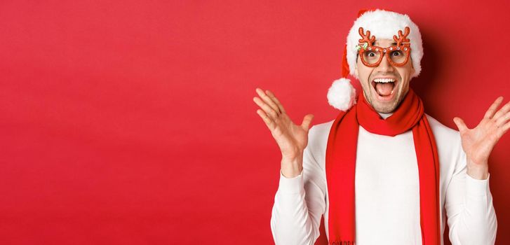 Concept of christmas, winter holidays and celebration. Image of surprised and happy man looking amazed, wearing party glasses and enjoying new year, standing over red background.