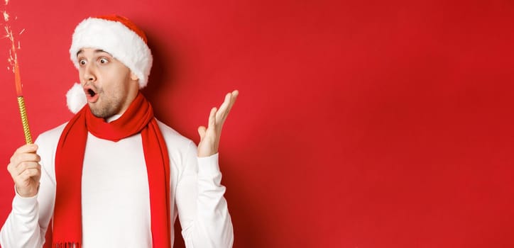 Concept of christmas, winter holidays and celebration. Close-up of amazed handsome man in santa hat and scarf, looking at sparkler with excitement, standing over red background.