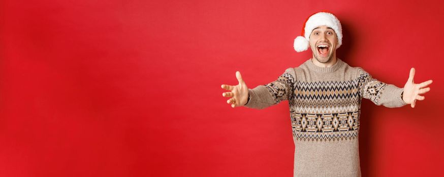 Portrait of happy, attractive man in christmas sweater and santa hat, reaching hands for gift, want to take something and smiling, standing over red background.