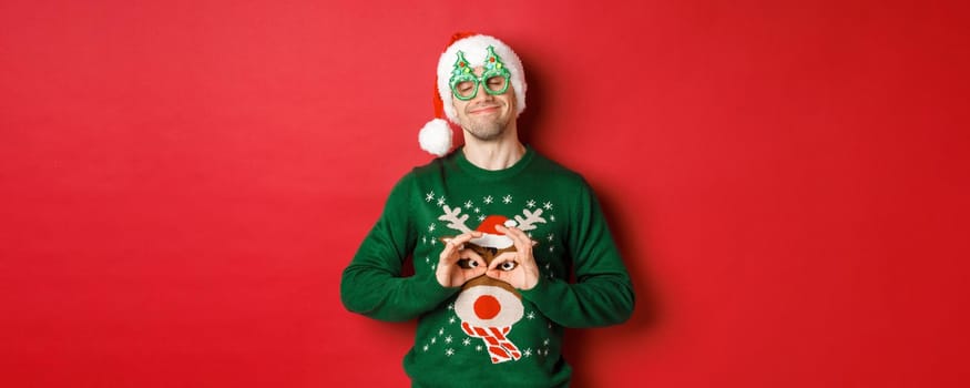 Image of happy smiling man in party glasses and santa hat, fooling around with funny christmas sweater, celebrating winter holidays, standing over red background.