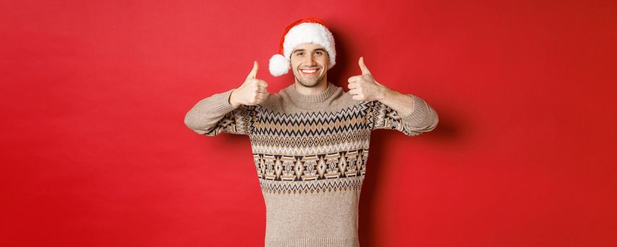 Portrait of attractive happy man in santa hat and sweater, showing thumbs-up in approval and smiling, wishing merry christmas, standing over red background.