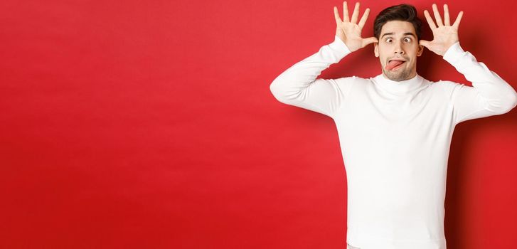 Portrait of funny caucasian guy, showing tongue and making faces, wearing white sweater, standing against red background.