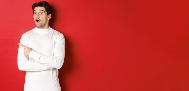 Concept of winter holidays. Portrait of impressed handsome man in white sweater, pointing and looking left with dropped jaw and amazed expression, standing against red background.