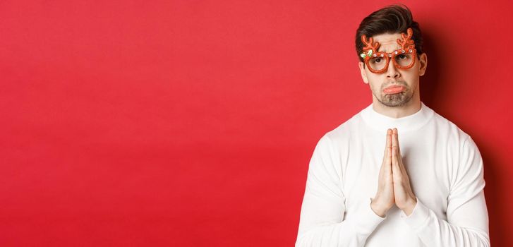 Image of handsome man in christmas party glasses, begging for help or apologizing, need a favour, standing over red background.