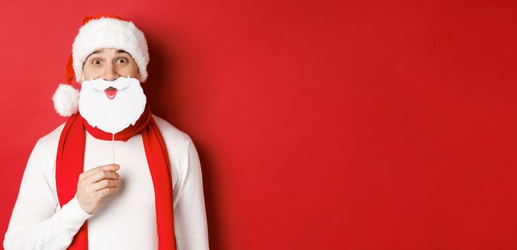 Concept of christmas, winter holidays and celebration. Portrait of funny man in santa hat, holding beard mask, enjoying new year party, standing over red background.