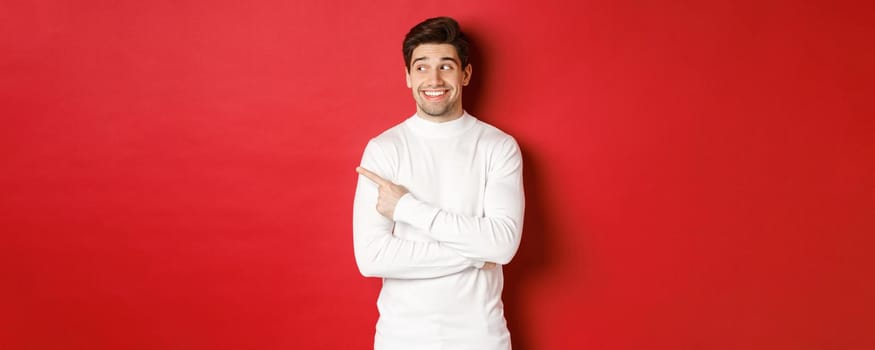 Concept of winter holidays, christmas and lifestyle. Excited handsome man in white sweater, smiling, looking and pointing finger left at promo offer, standing over red background.