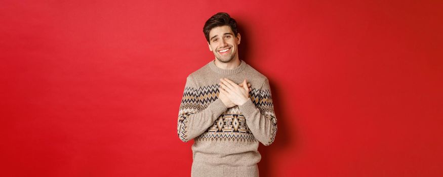 Portrait of touched and happy handsome guy, receiving new year gift, holding hands on heart and smiling, saying thank you, standing in christmas sweater against red background.