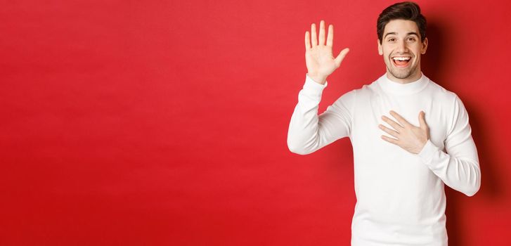 Portrait of honest smiling man in white sweater, making a promise, swearing to tell truth, standing against red background.