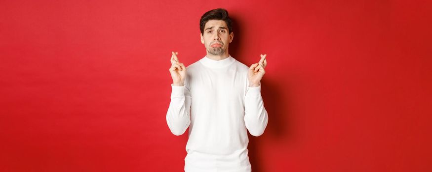 Image of sad and gloomy guy in white sweater, crying with fingers crossed, waiting for something or praying, standing over red background.